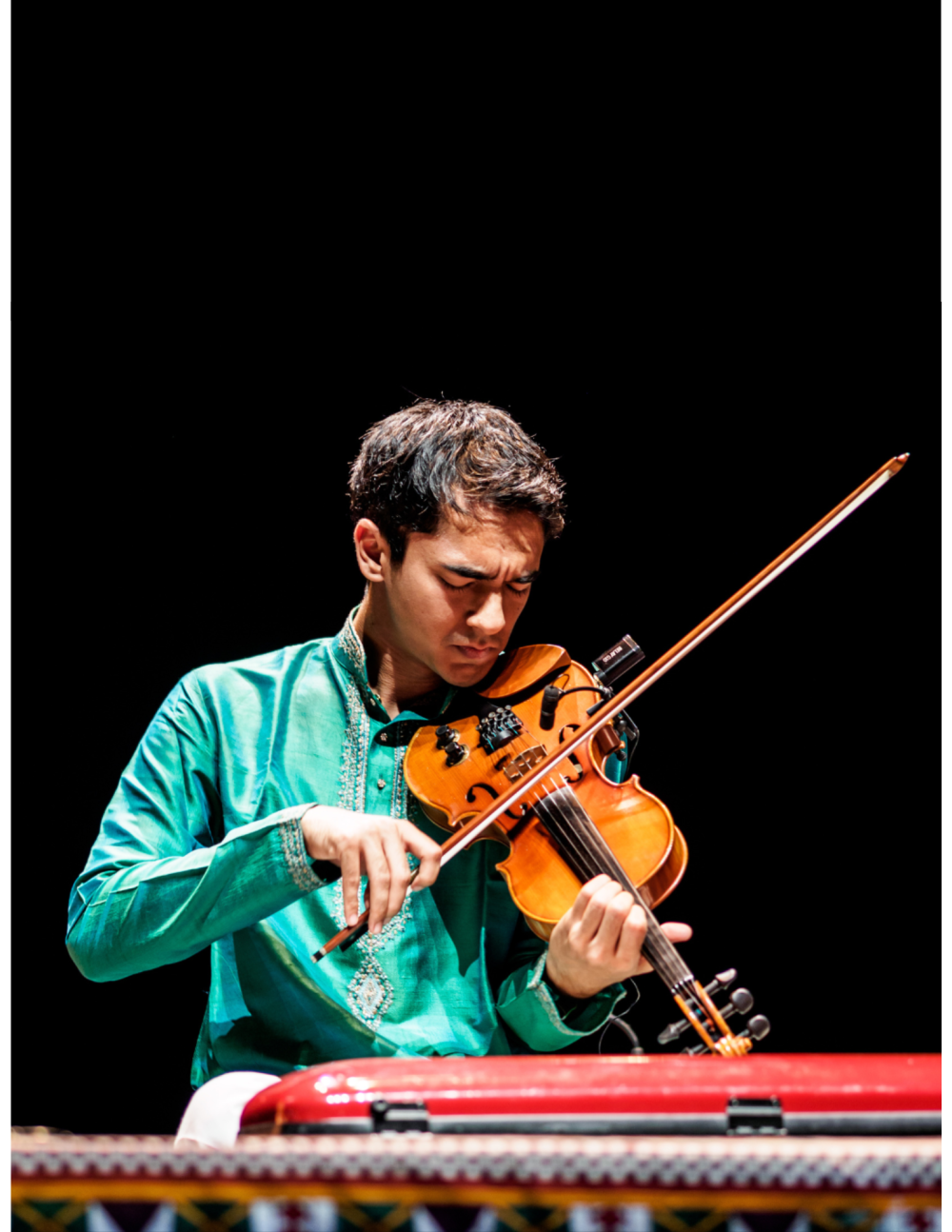 Ambi Subramaniam is sitting on stage playing violin in a green tunic. 
